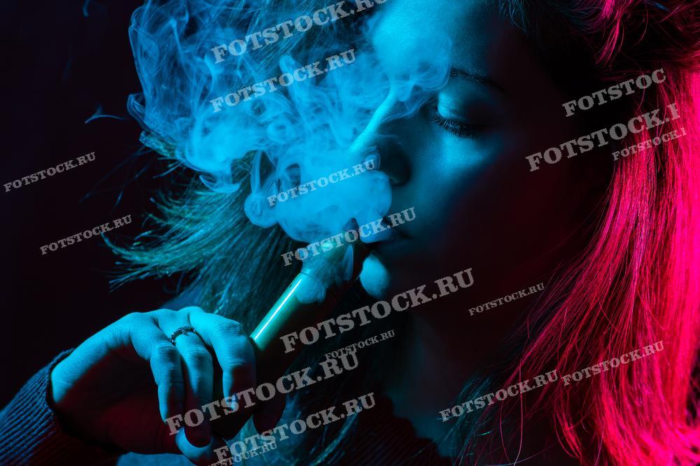 Фотография изображает молодую женщину, выпускающую облако пара от электронной сигареты в неоновом свете.